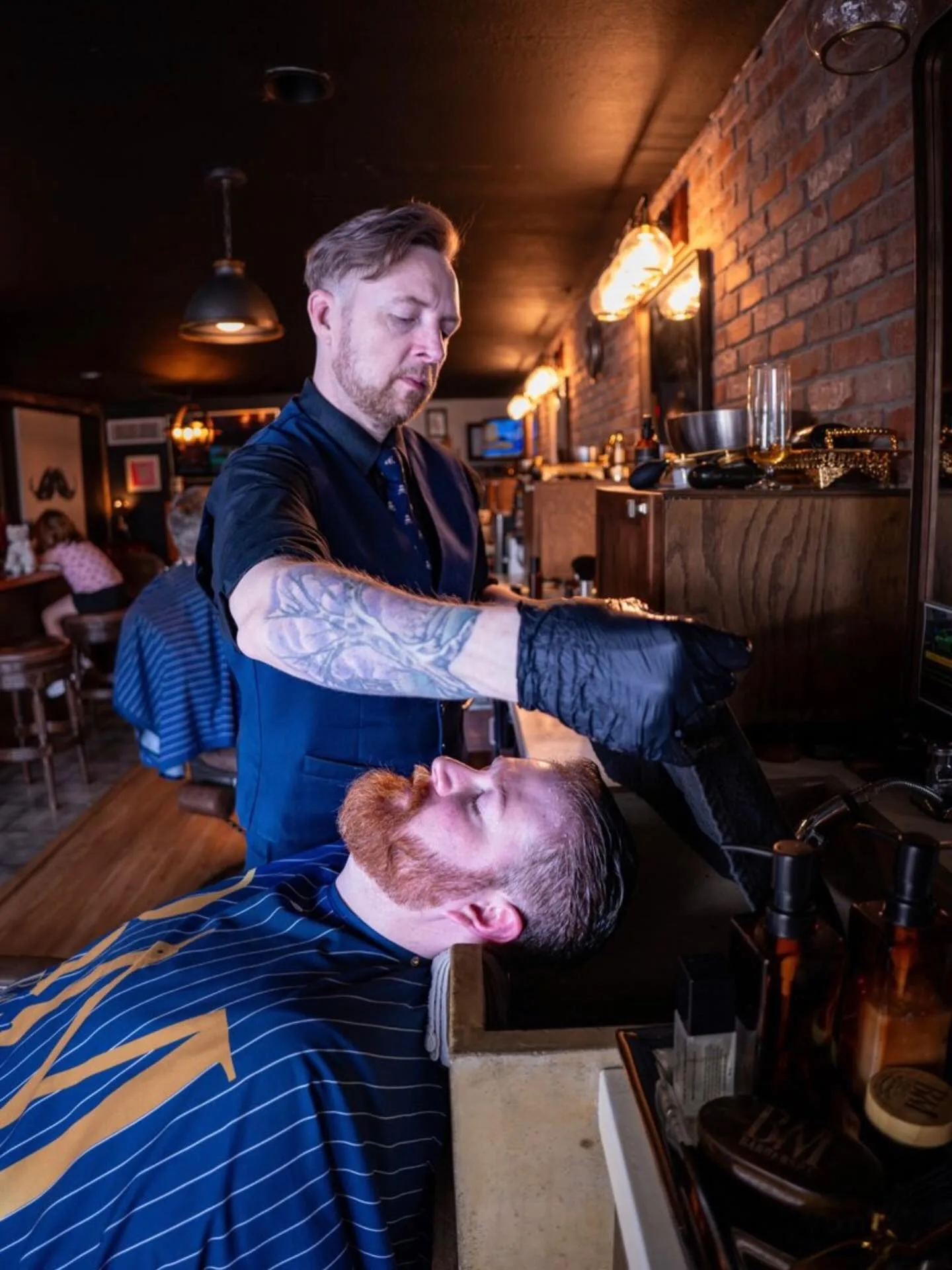 A Barley and Mane barber in a navy vest giving a reclined client a hot towel beard treatment under warm Edison bulb lighting