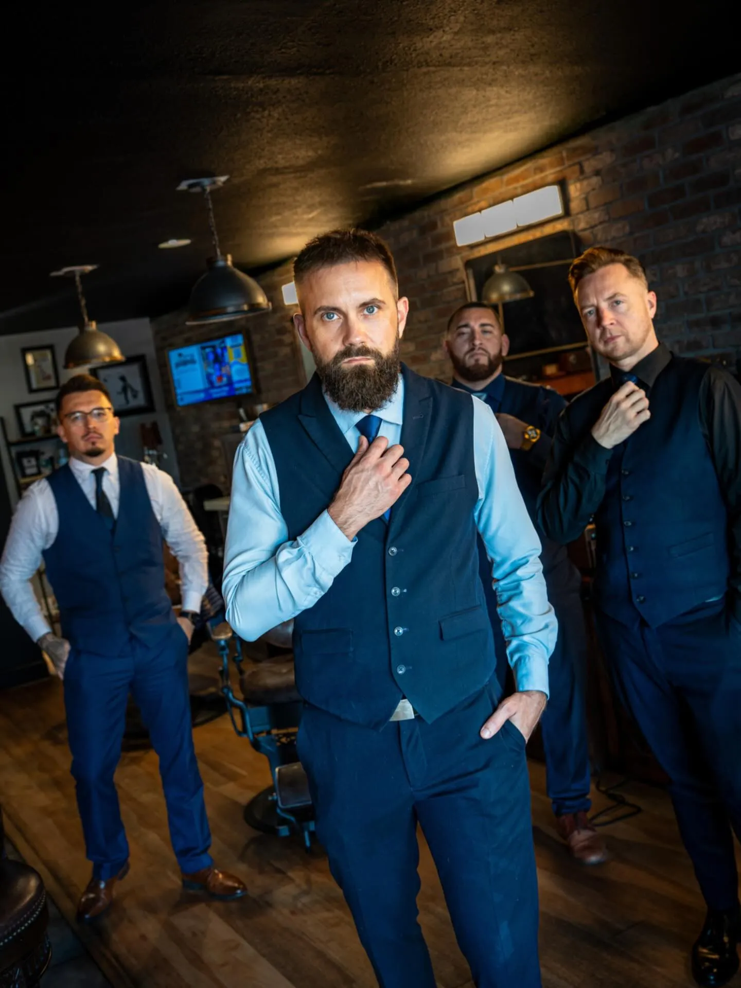 Four Barley and Mane barbers in navy vests straightening their ties on the shop floor