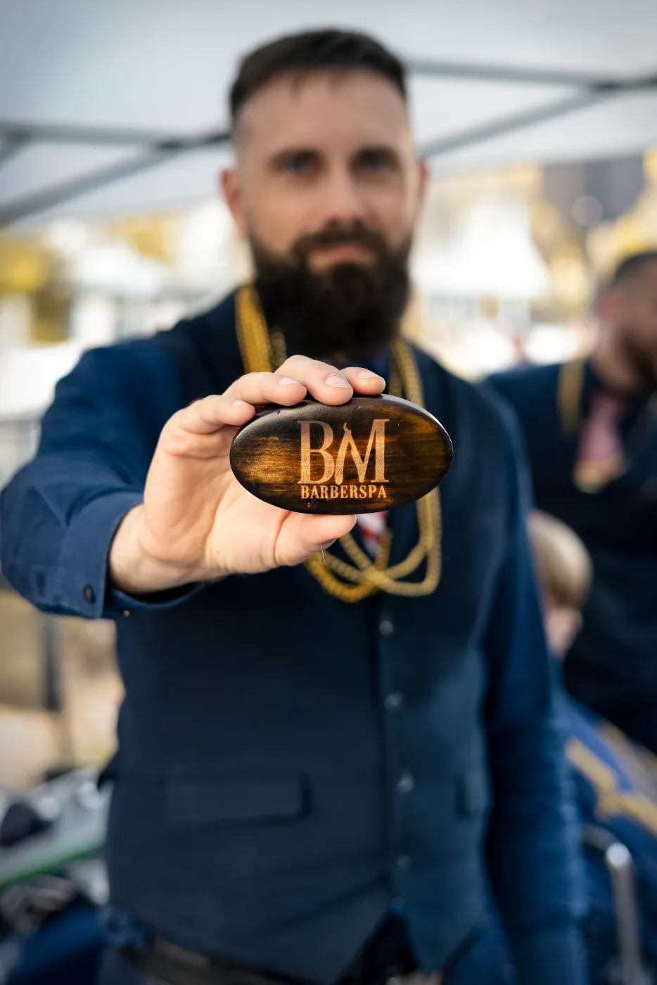 A bearded barber in a navy vest holding a wooden Barley and Mane BarberSpa beard brush toward the camera