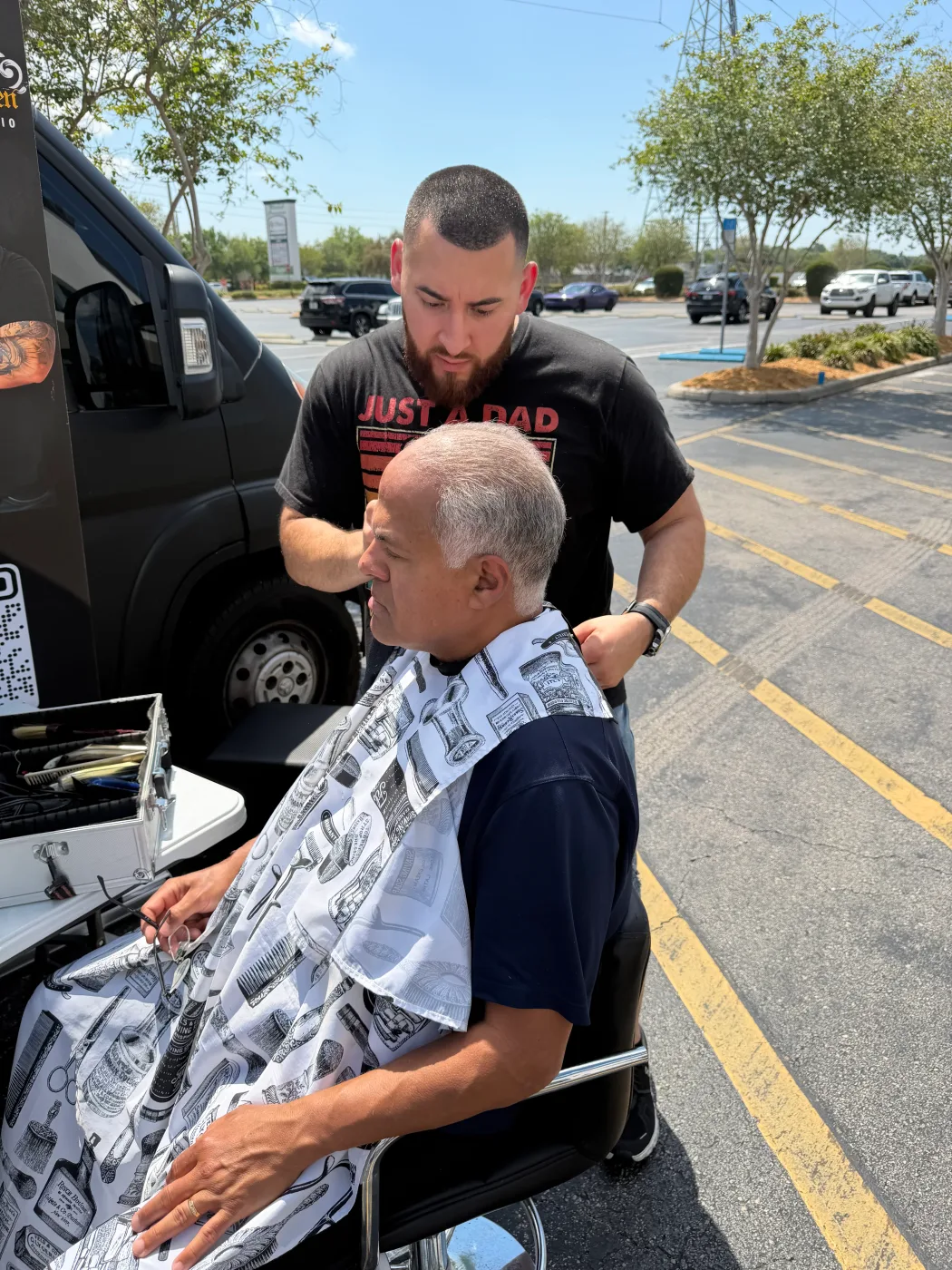 A Barley and Mane barber cutting a client's hair in a barber chair set up outdoors beside a van