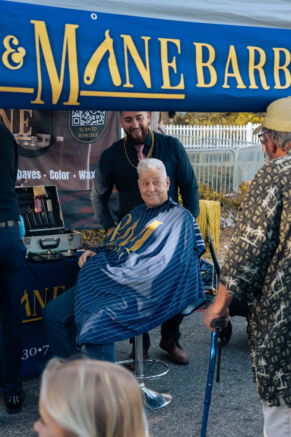 A smiling older client in a Barley and Mane cape in the chair, with his barber standing behind him at an outdoor event