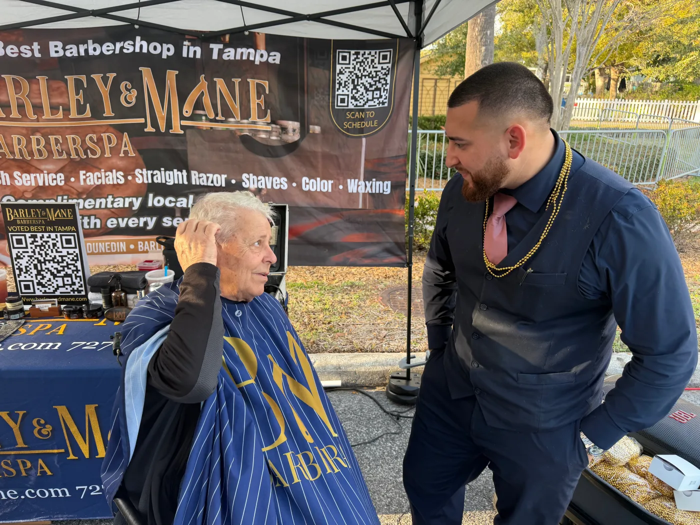 A Barley and Mane barber talking with a seated client in front of the shop banner at an outdoor event