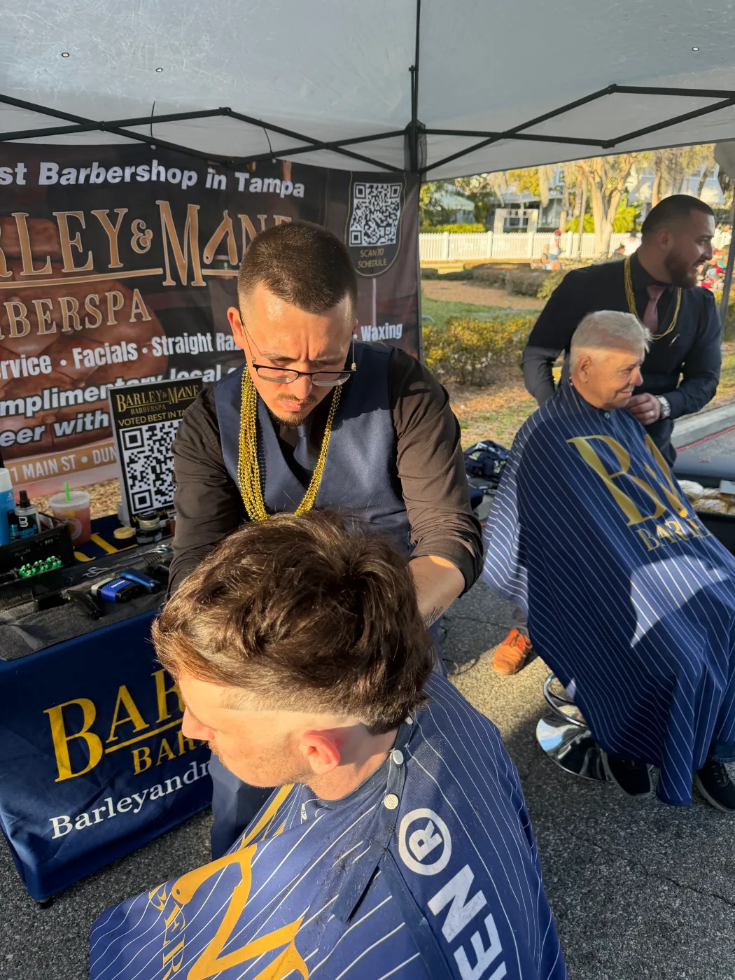 Two Barley and Mane barbers cutting hair under the shop canopy at an outdoor event in Dunedin