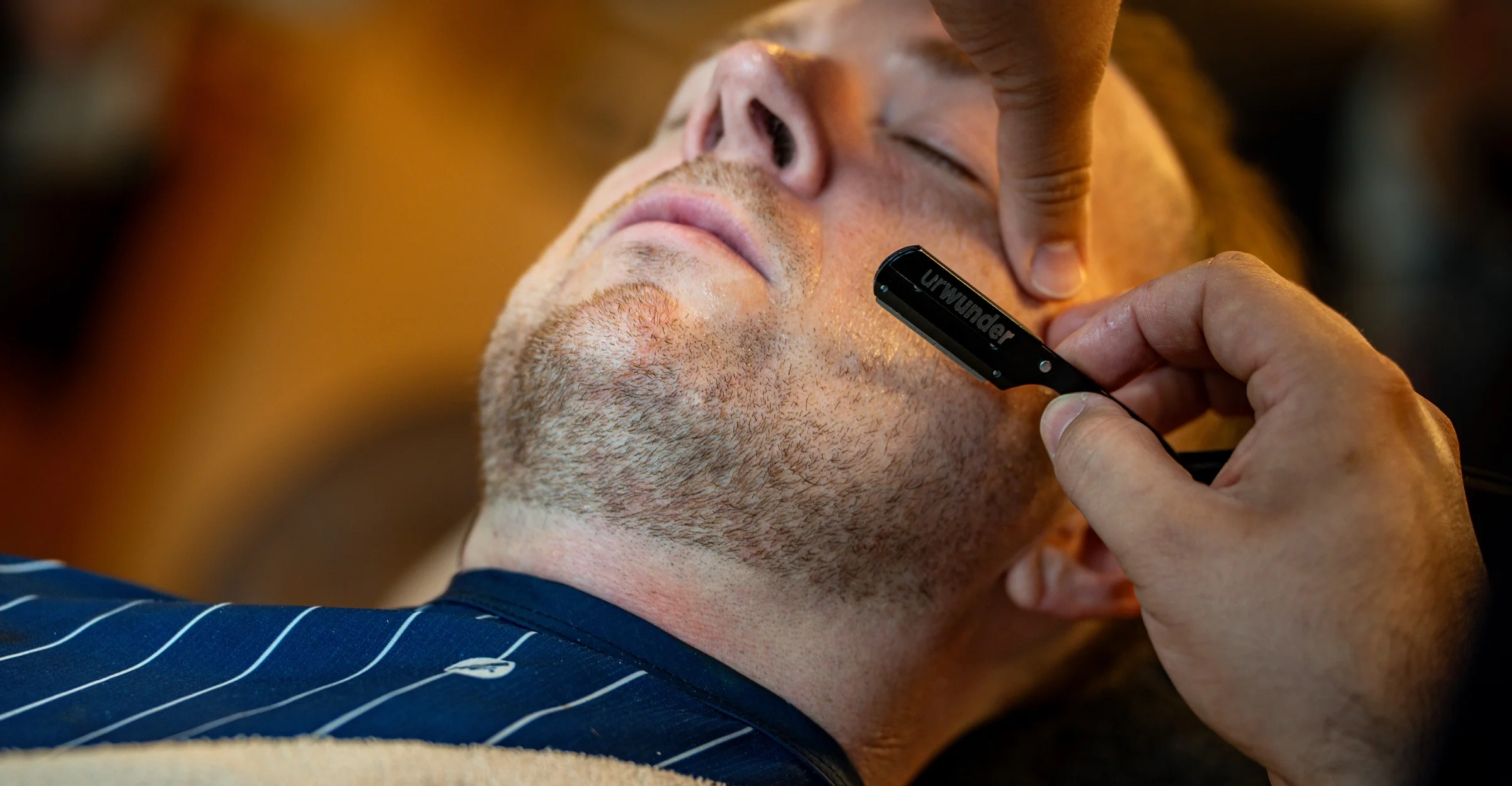 A barber shaving a reclined client's jaw with a straight razor