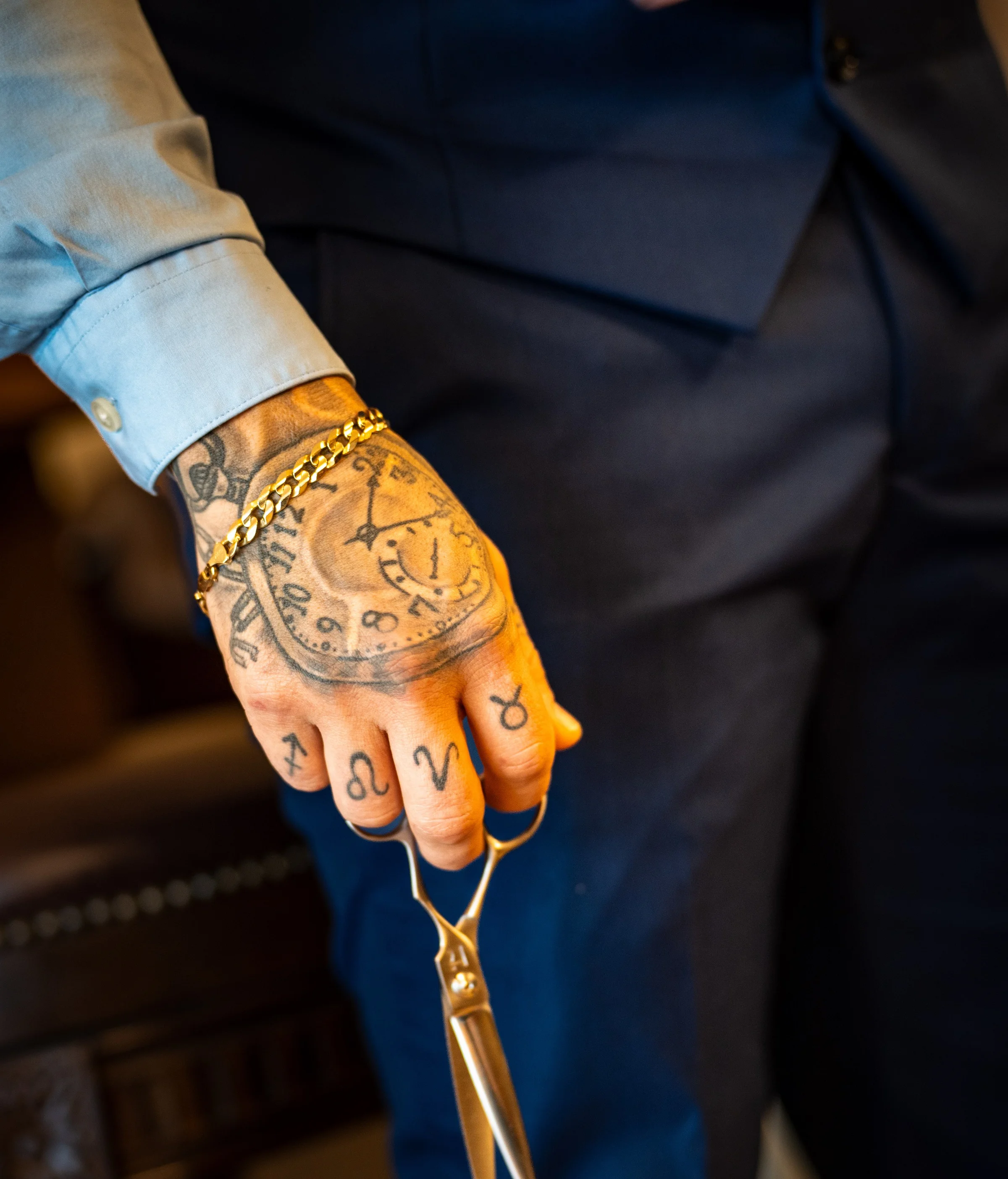 A barber's tattooed hand holding a pair of shears, a gold chain at his wrist