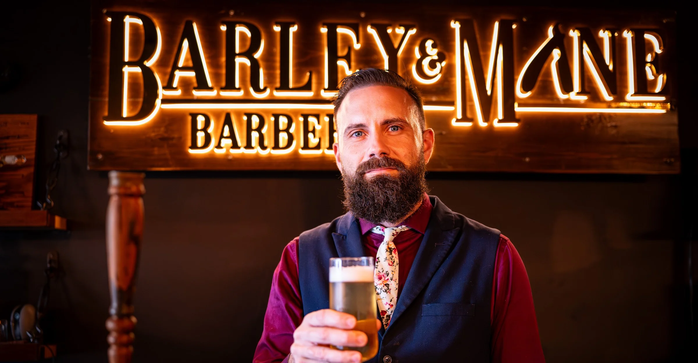 Owner Ryan in a navy vest raising a glass of beer beneath the lit Barley and Mane sign