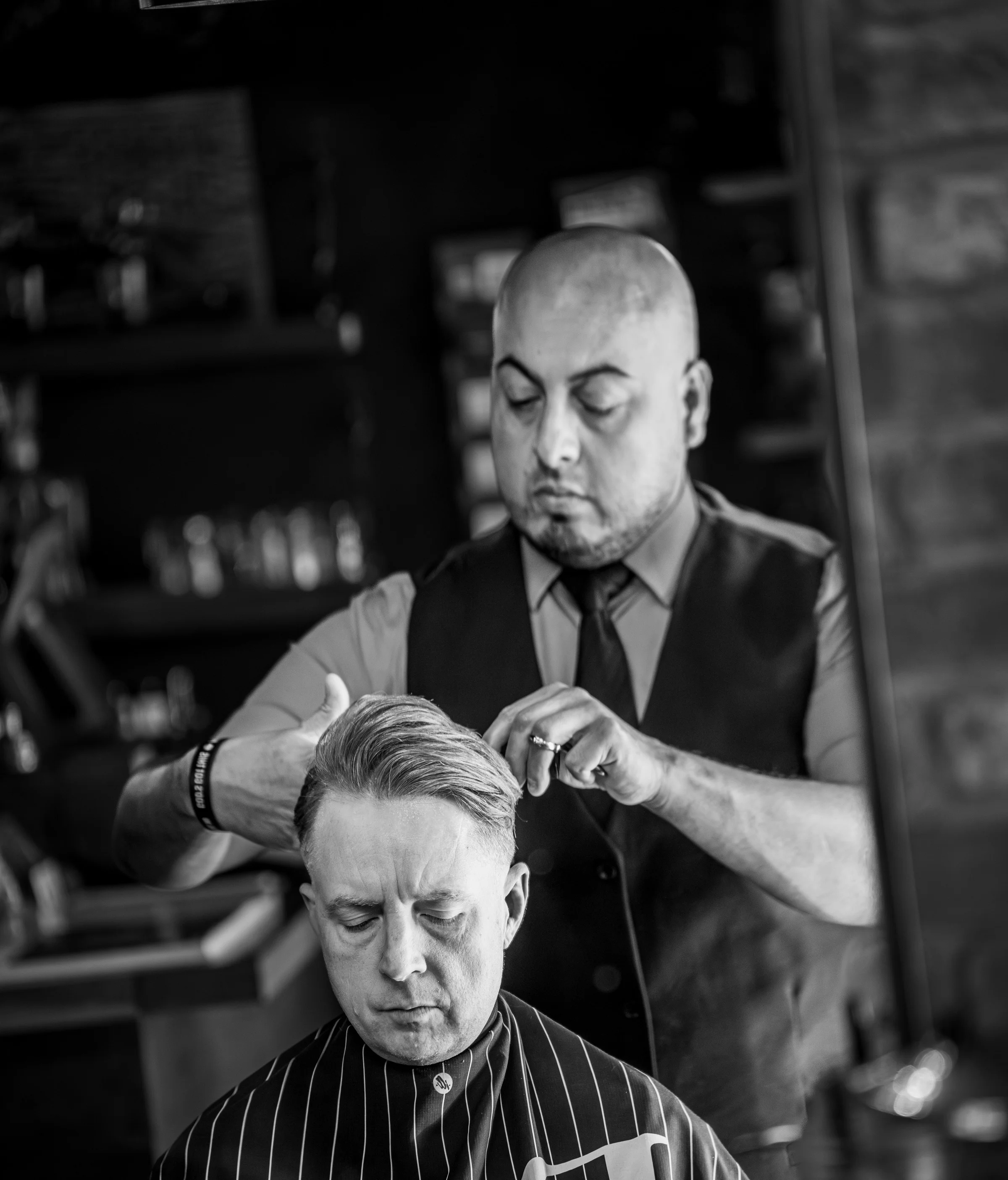 A black and white photo of a Barley and Mane barber in a vest and tie finishing a client's haircut