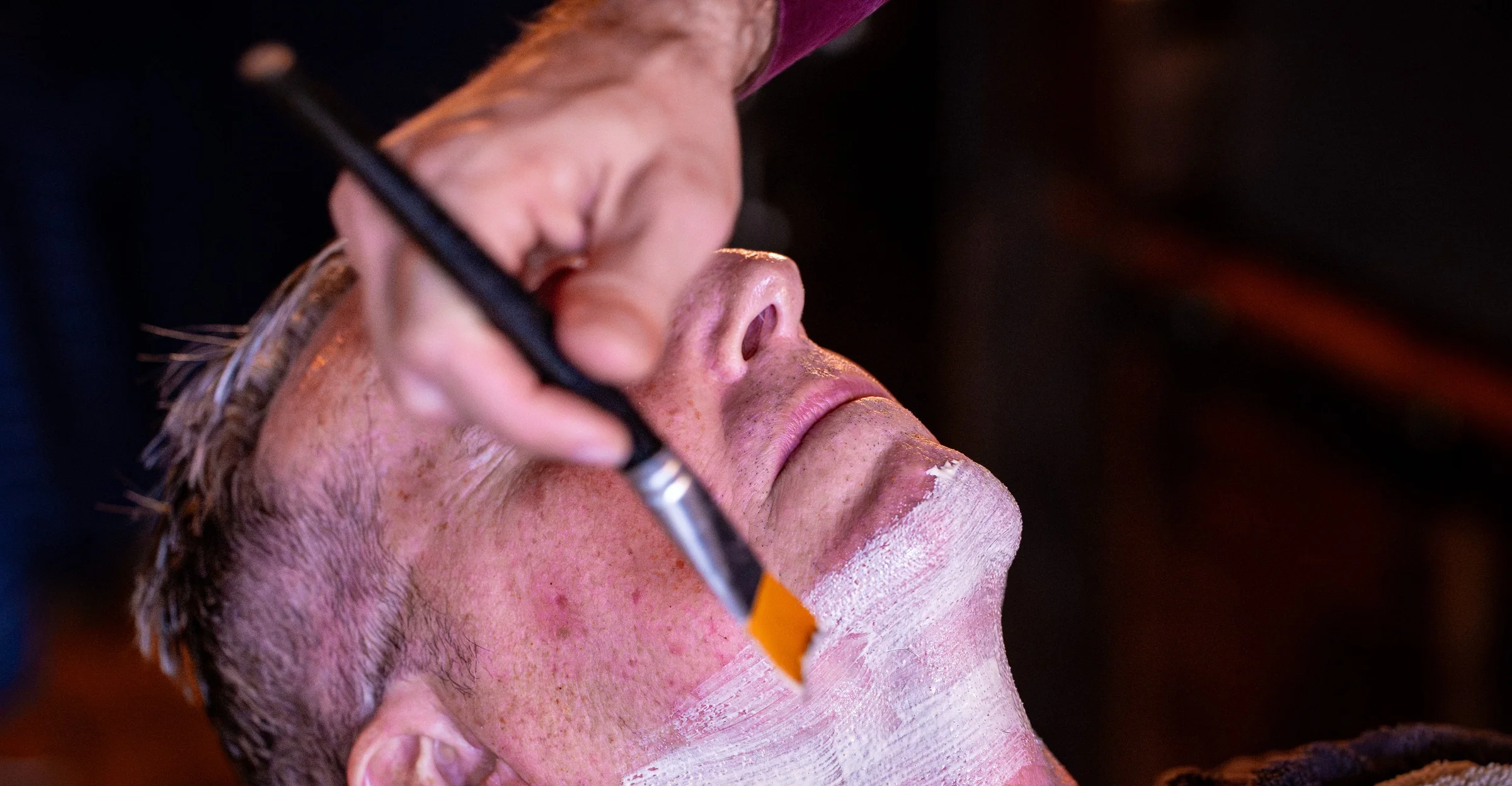 A client reclined with his eyes closed while a barber brushes a clay mask onto his face