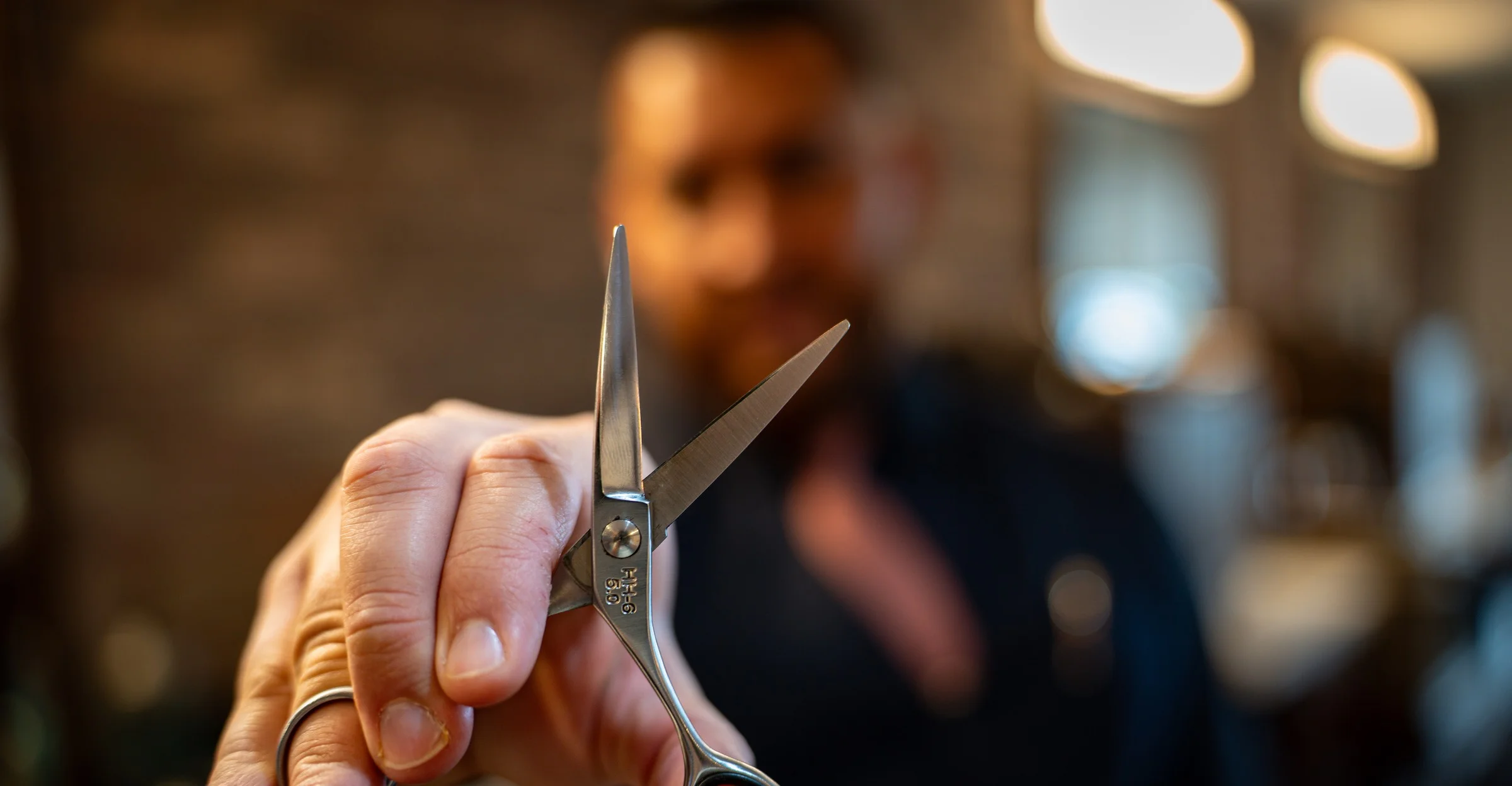 A barber holding a pair of shears toward the camera, the shop warm and out of focus behind him