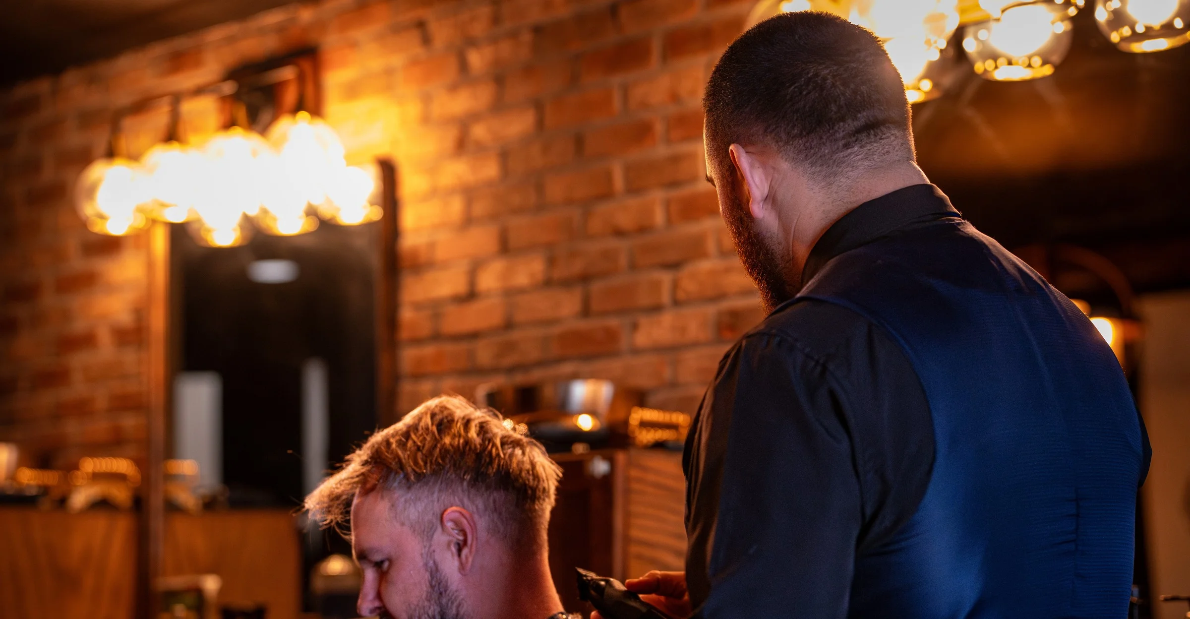 A Barley and Mane barber in a vest cutting a client's hair under Edison bulbs against the shop's brick wall