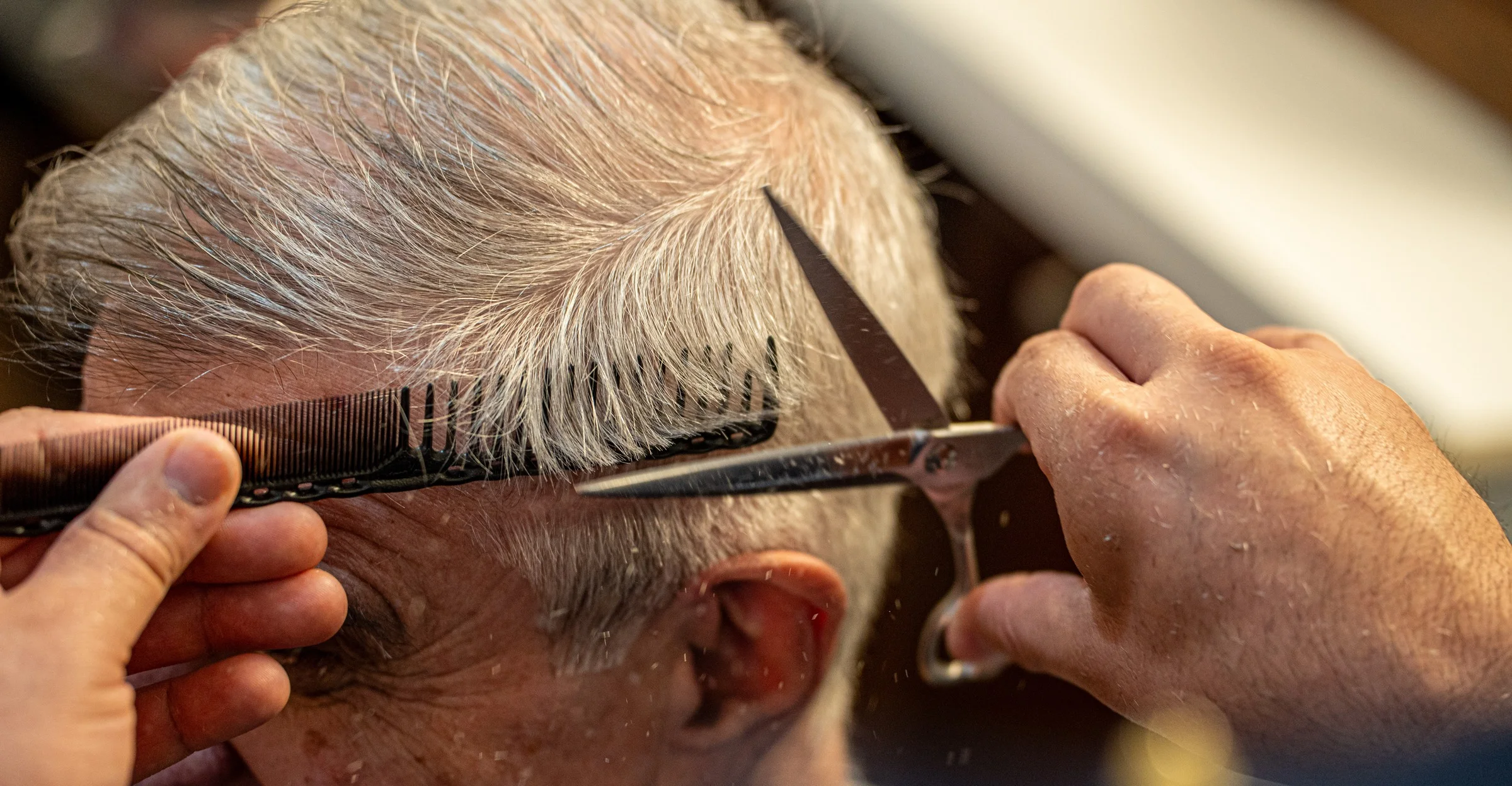A barber cutting a client's grey hair with scissors over a comb