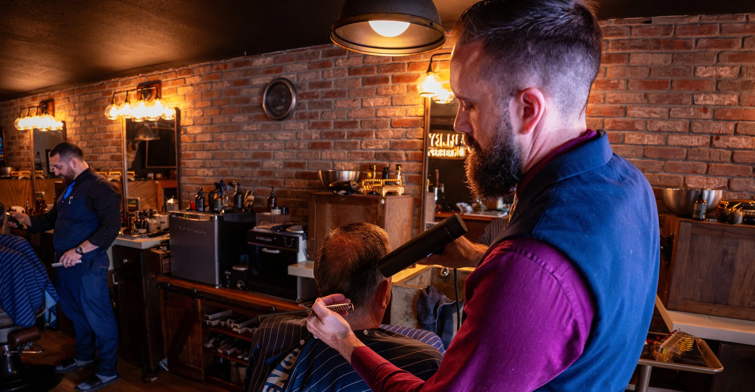 A Barley and Mane barber in a navy vest cutting a client's hair in the brick-walled shop under warm lights