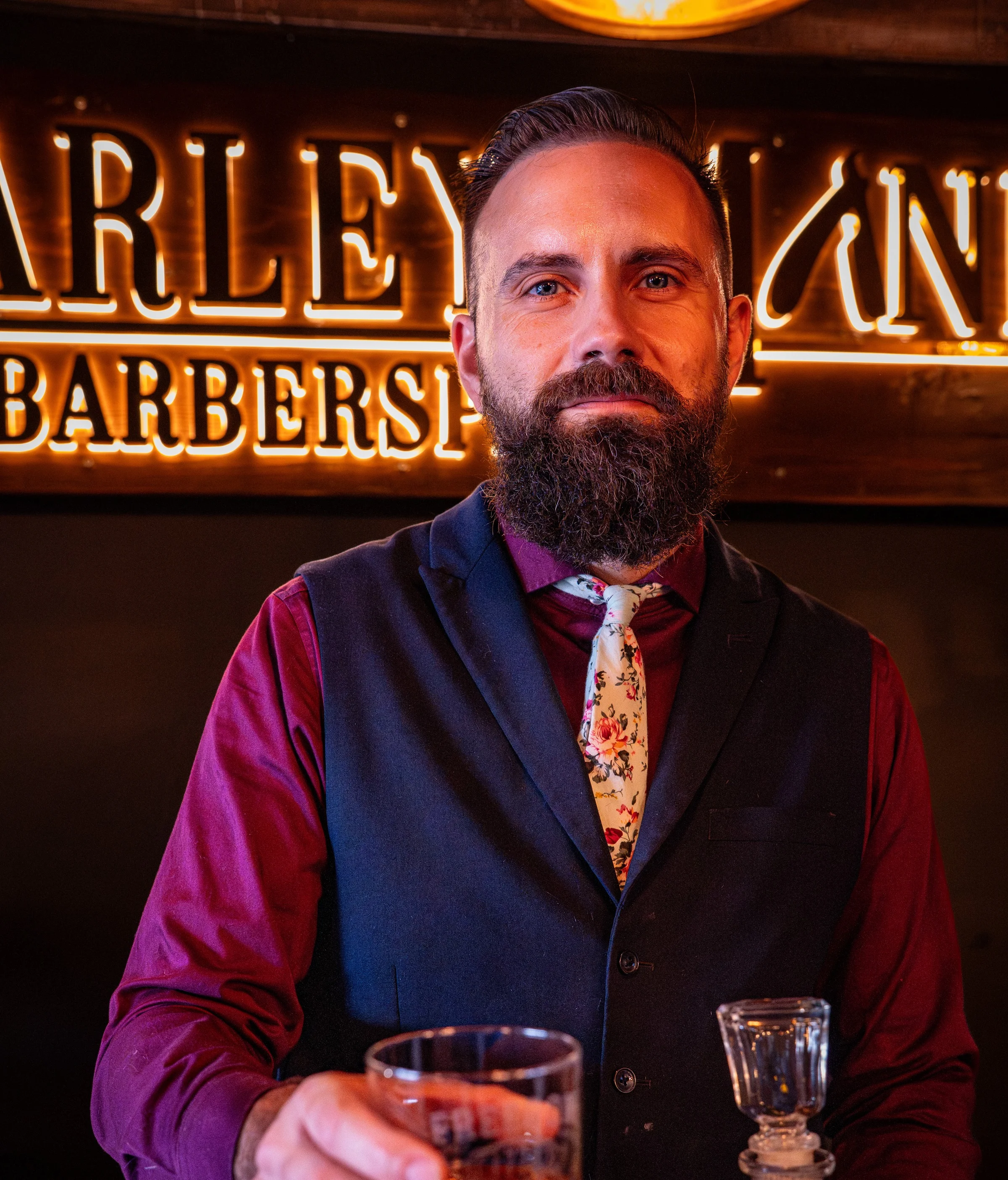 Owner Ryan in a navy vest and floral tie beneath the lit Barley and Mane sign