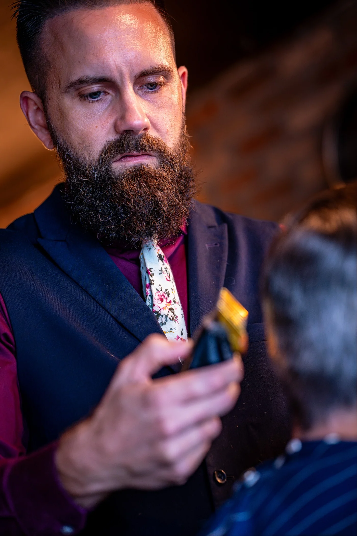 A bearded barber in a floral tie trimming a client's beard with gold clippers