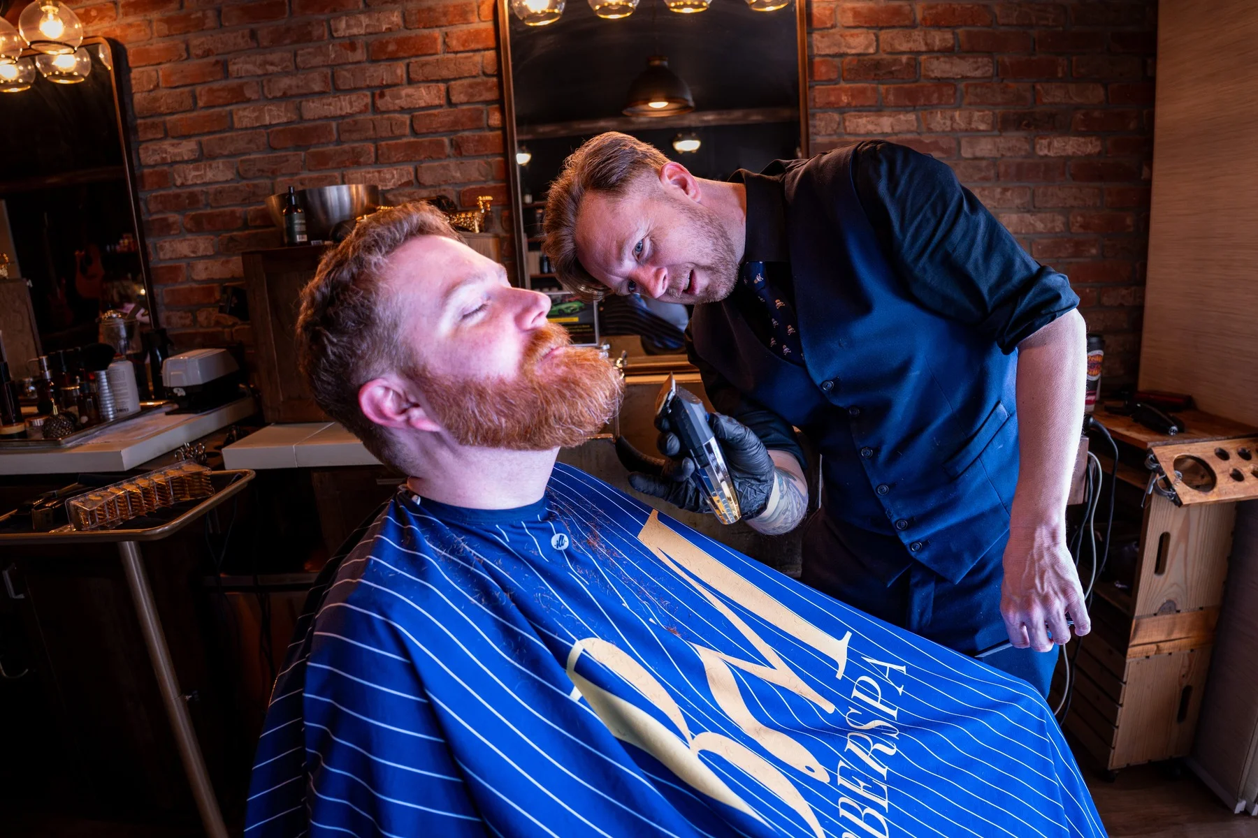 A barber in a vest trimming a relaxed client's red beard, the client wrapped in a Barley and Mane cape