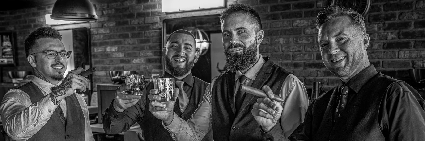 Four Barley and Mane barbers in vests raising glasses of whiskey and cigars, in black and white