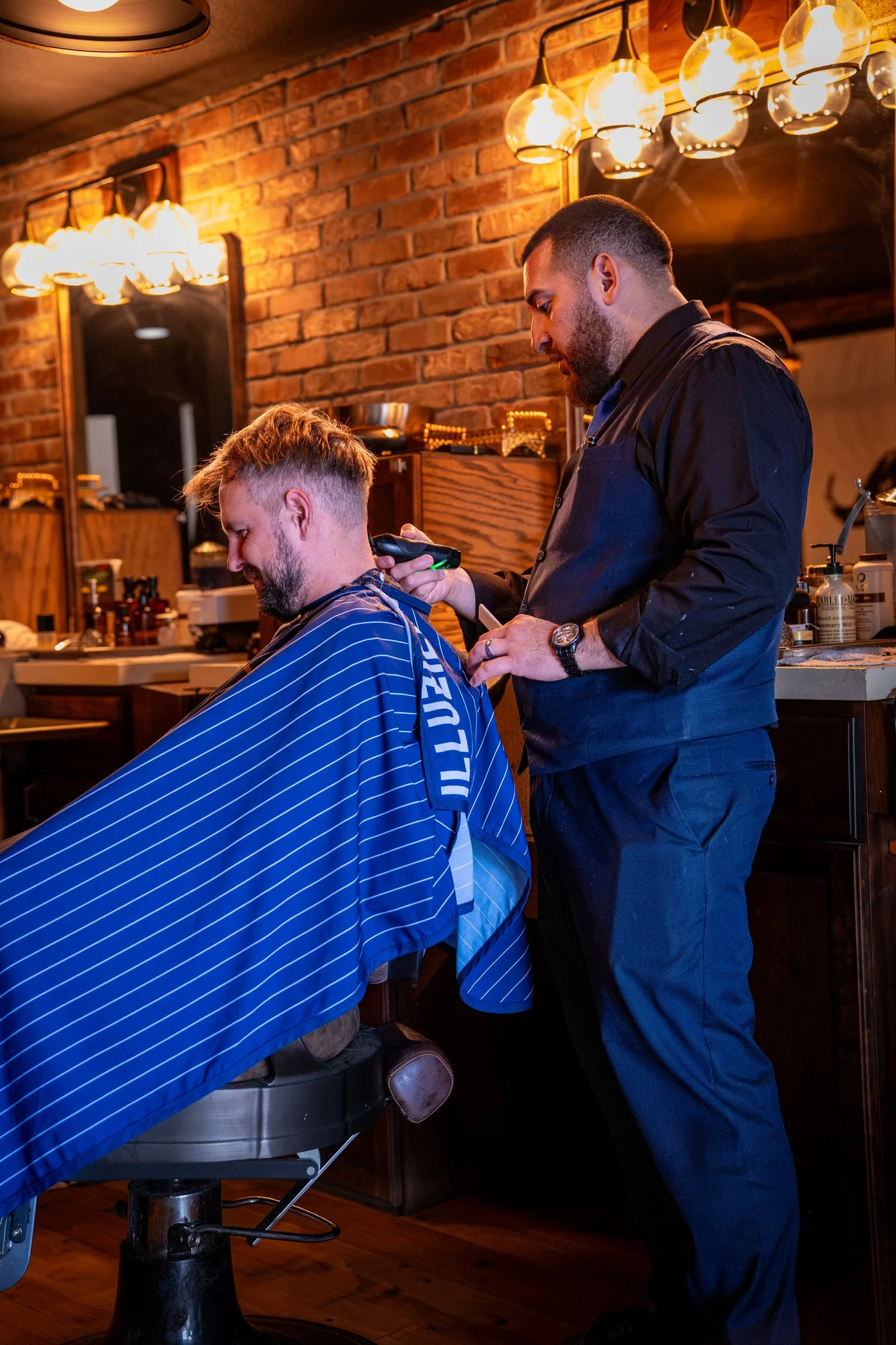 A Barley and Mane barber cutting a client's hair in a striped cape under a row of Edison bulbs