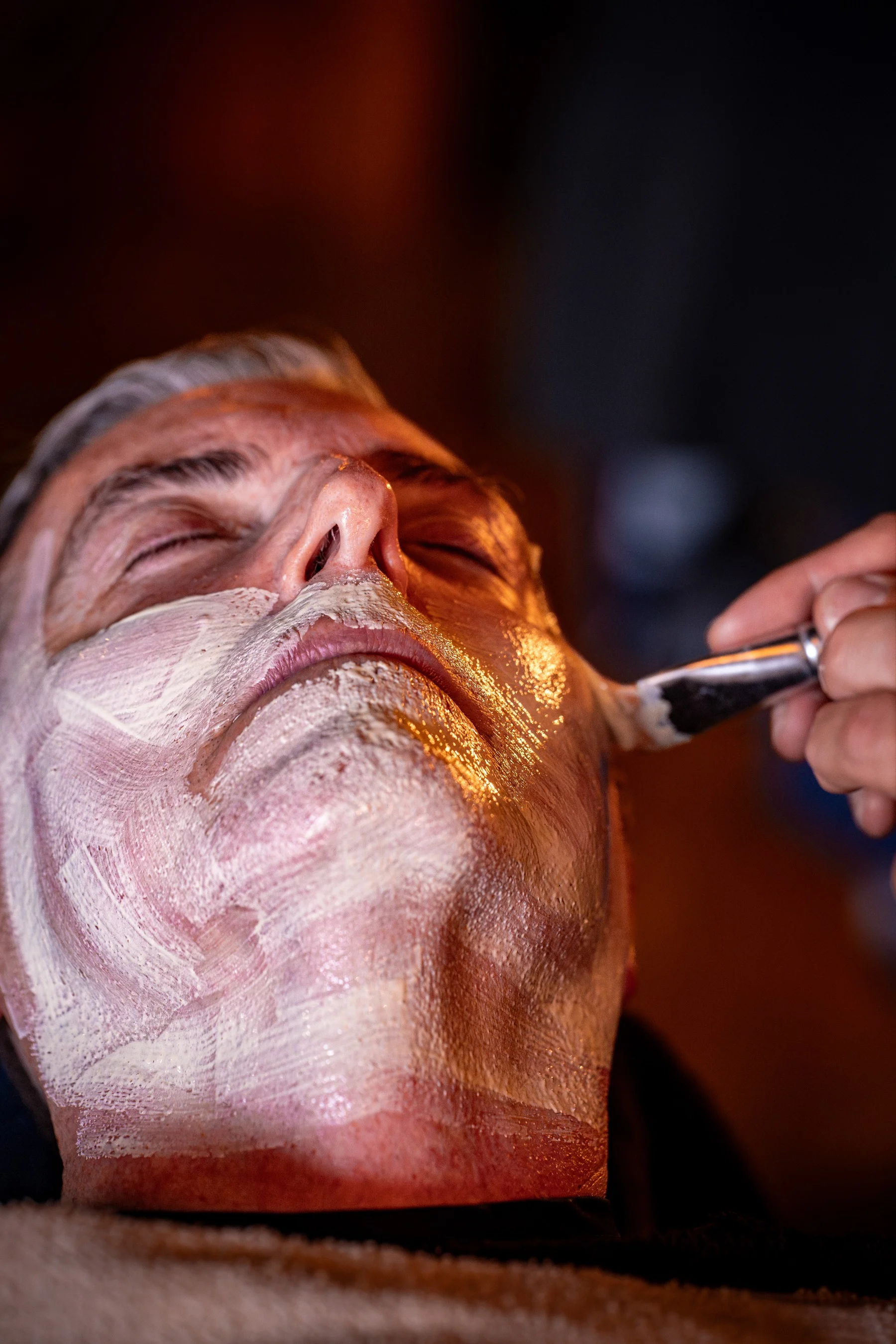 A brush spreading a mask across a reclined client's face during a men's facial