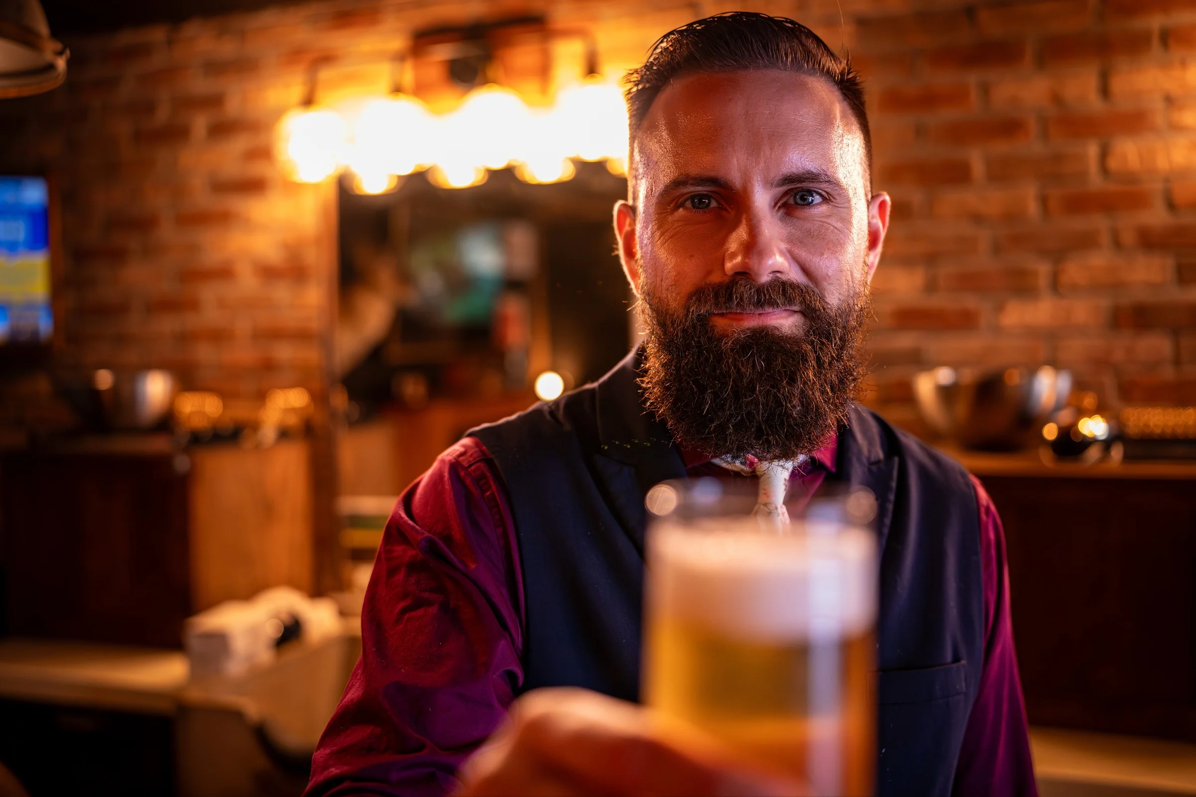 A bearded Barley and Mane barber smiling and holding out a freshly poured beer under warm Edison bulbs