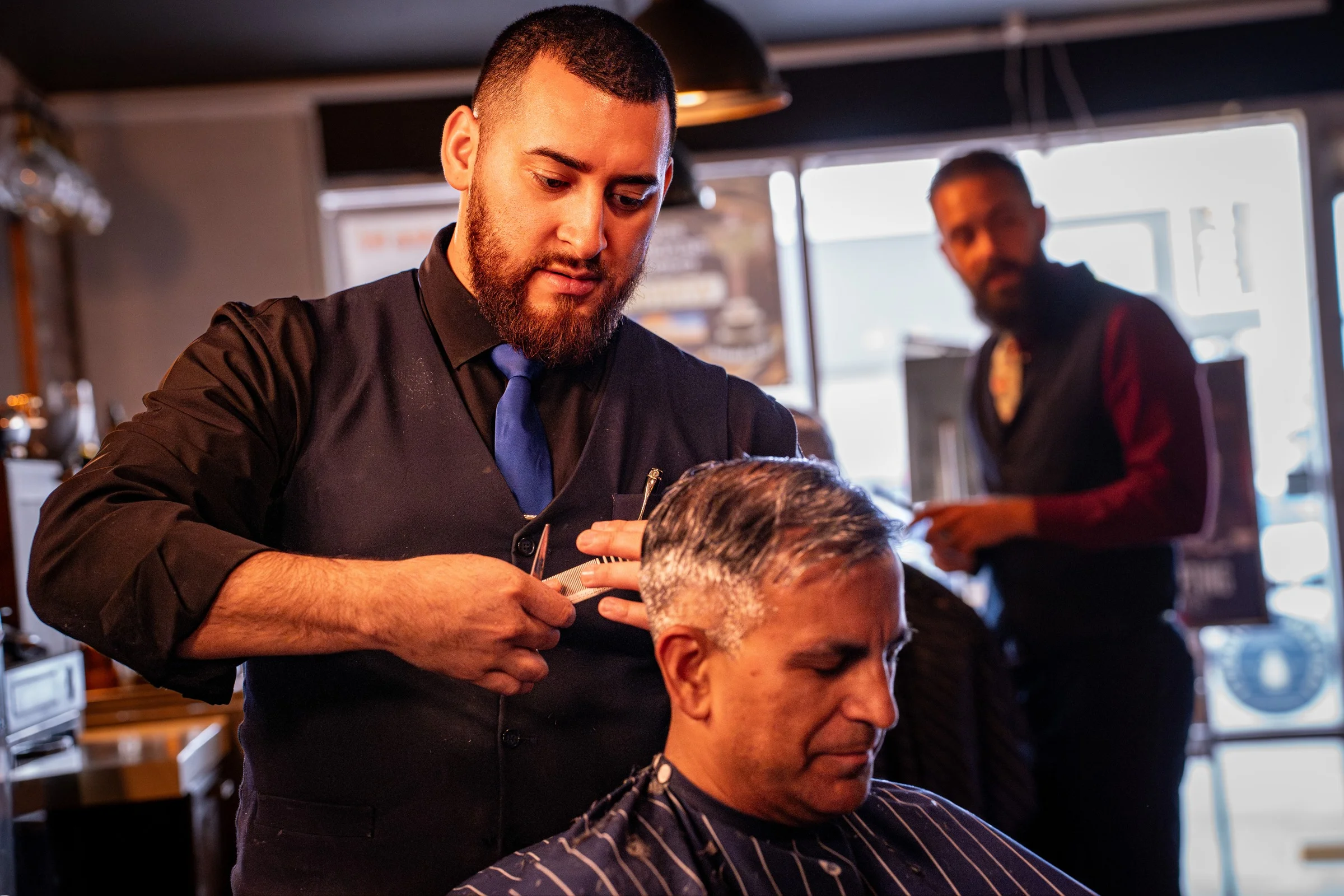 A Barley and Mane barber cutting a gray haired client's hair while another barber works by the front window