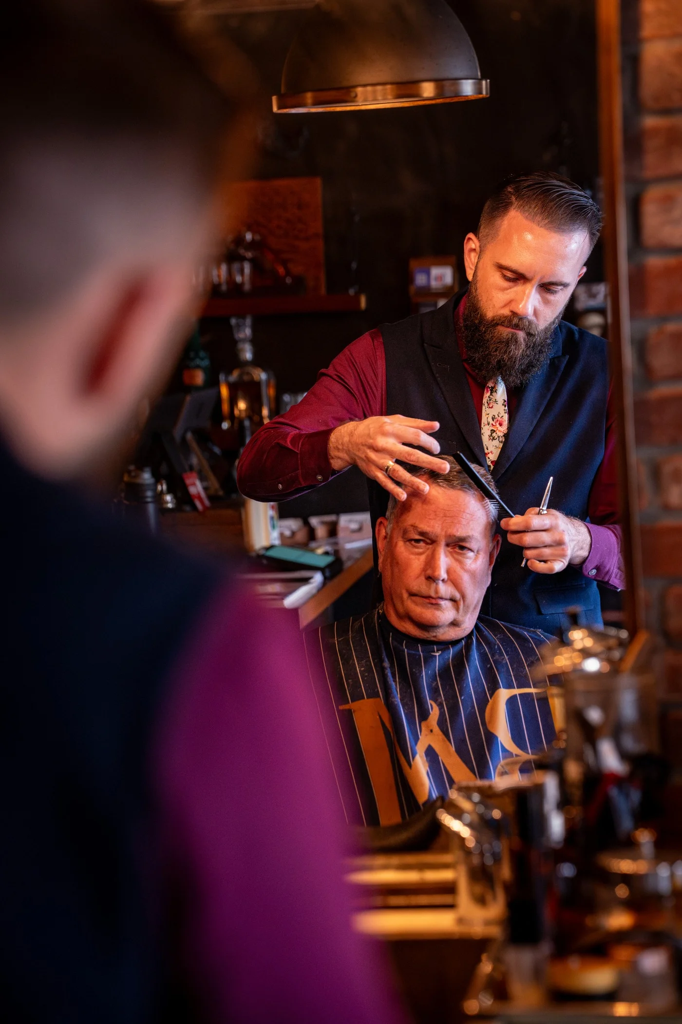 A bearded Barley and Mane barber cutting an older client's gray hair, seen past another client in the foreground