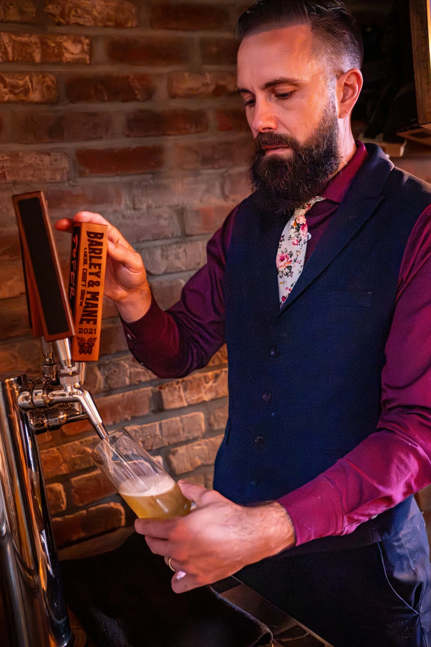 A Barley and Mane barber in a vest and floral tie pulling a beer from the tap against the brick wall