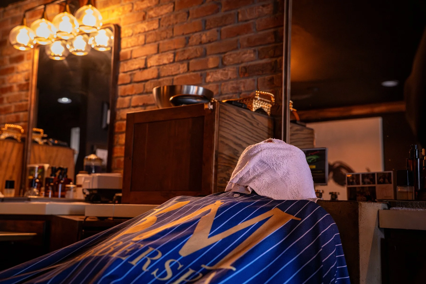 A client in a Barley and Mane cape with a towel wrapped over his head at a barber station under Edison bulbs