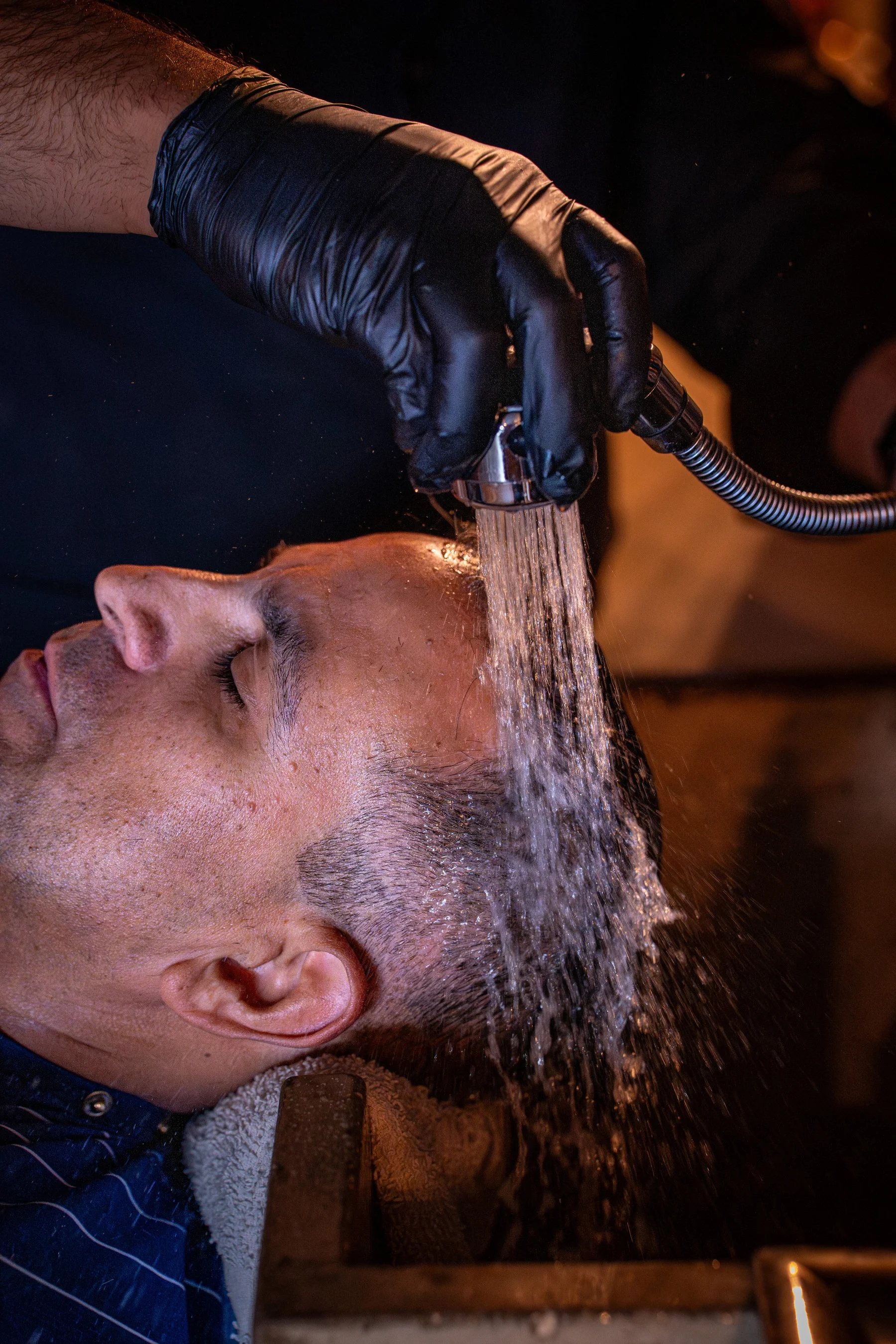 A barber in black gloves rinsing a reclined client's hair with a sprayer at the shampoo bowl