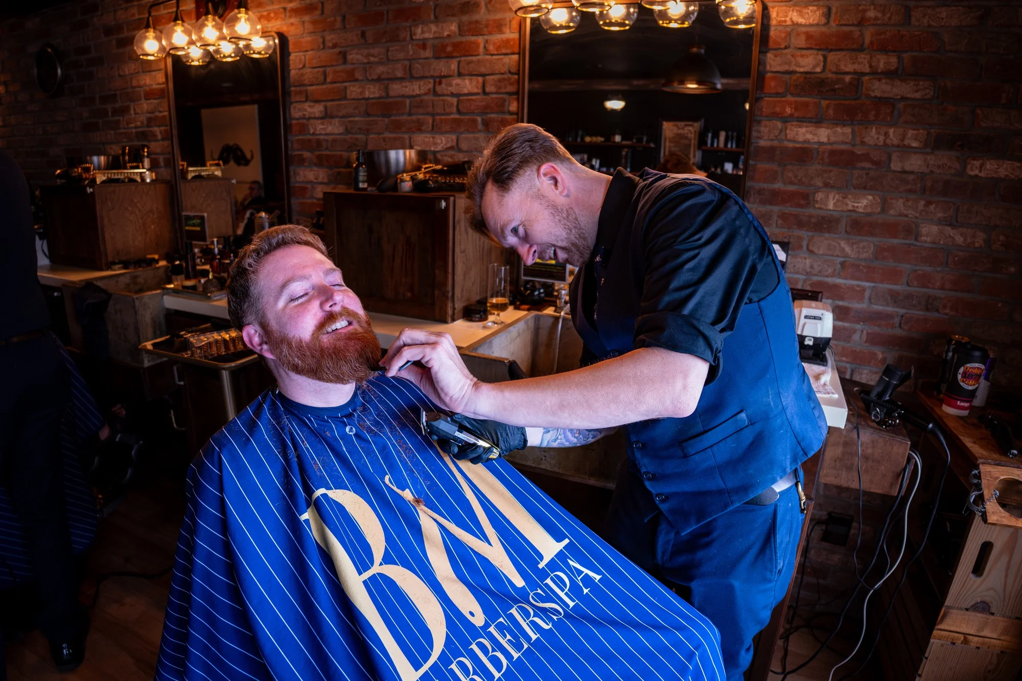 A barber trimming a laughing client's red beard, the client wrapped in a Barley and Mane cape