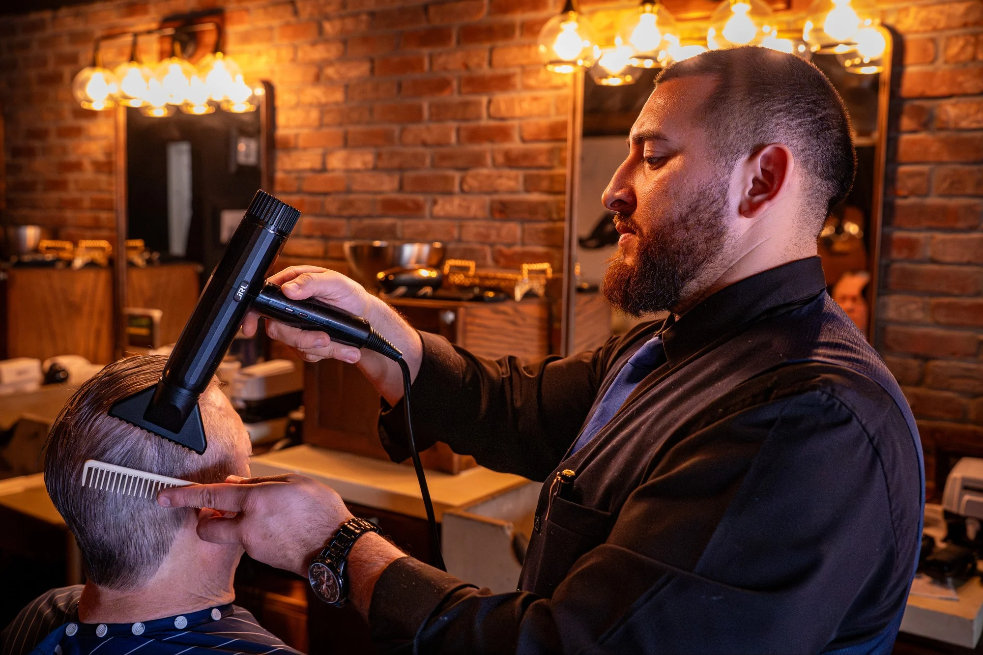 A Barley and Mane barber blow drying and combing a client's hair under a row of Edison bulbs