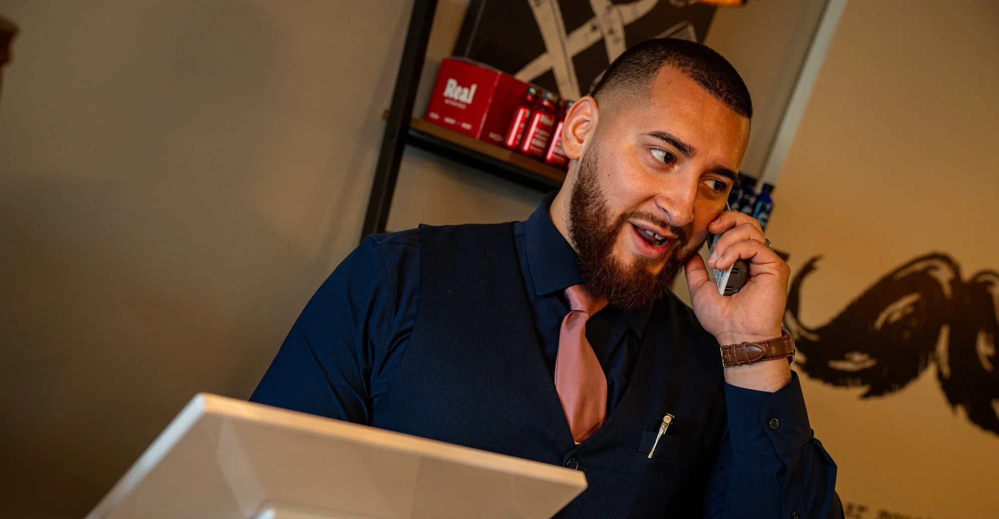 A Barley and Mane barber in a vest and tie taking a call at the front desk
