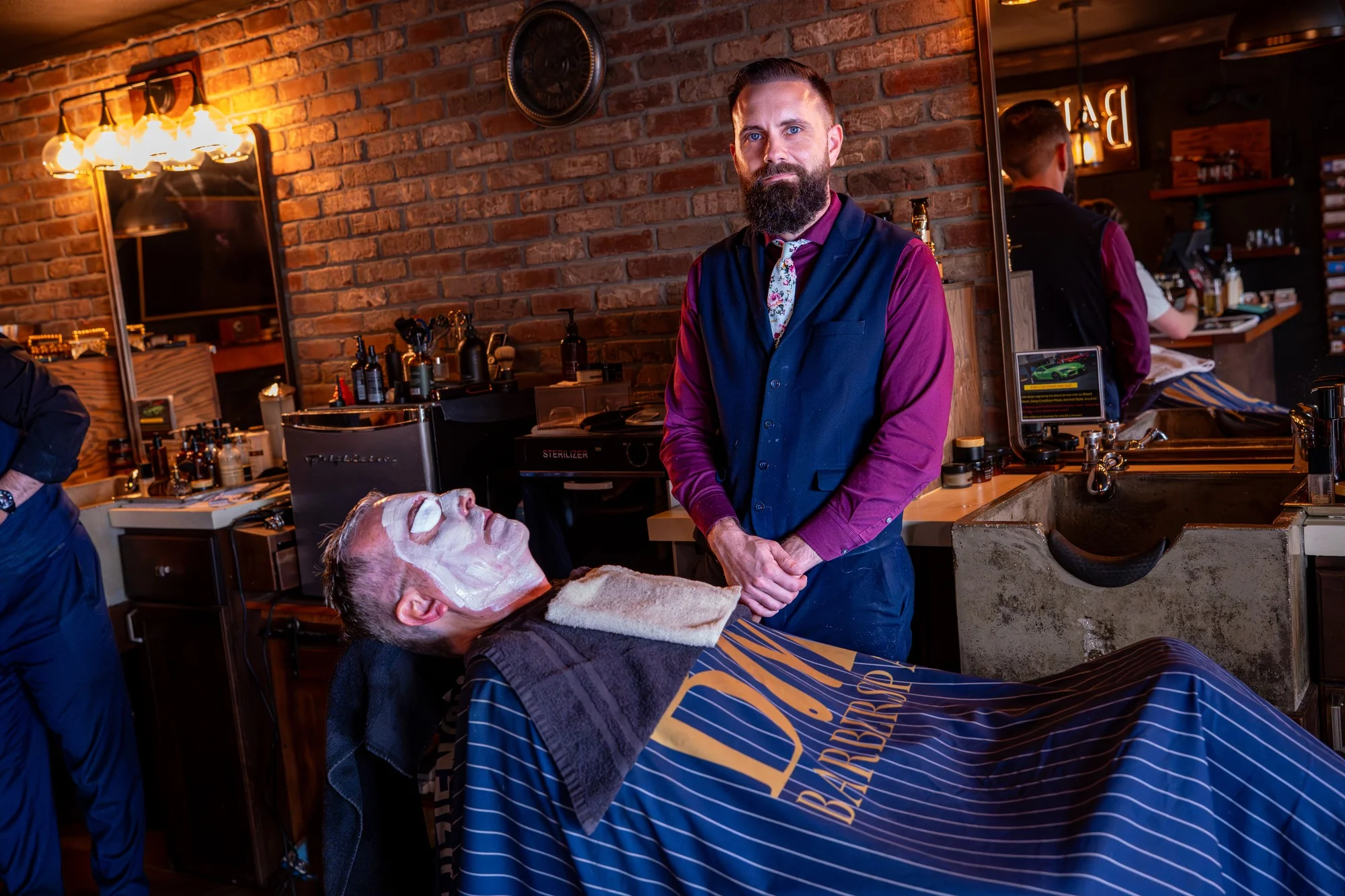 Owner Ryan standing beside a client relaxing under a facial mask in the Barley and Mane chair