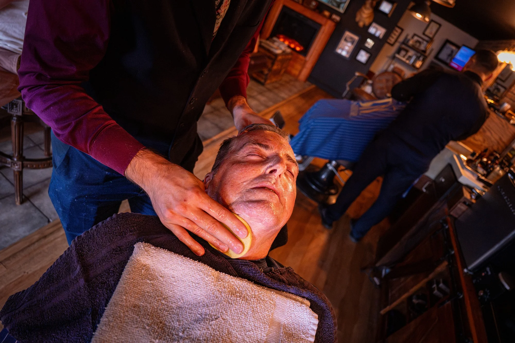 A barber tending to a reclined client wrapped in a towel, with the shop floor busy behind them